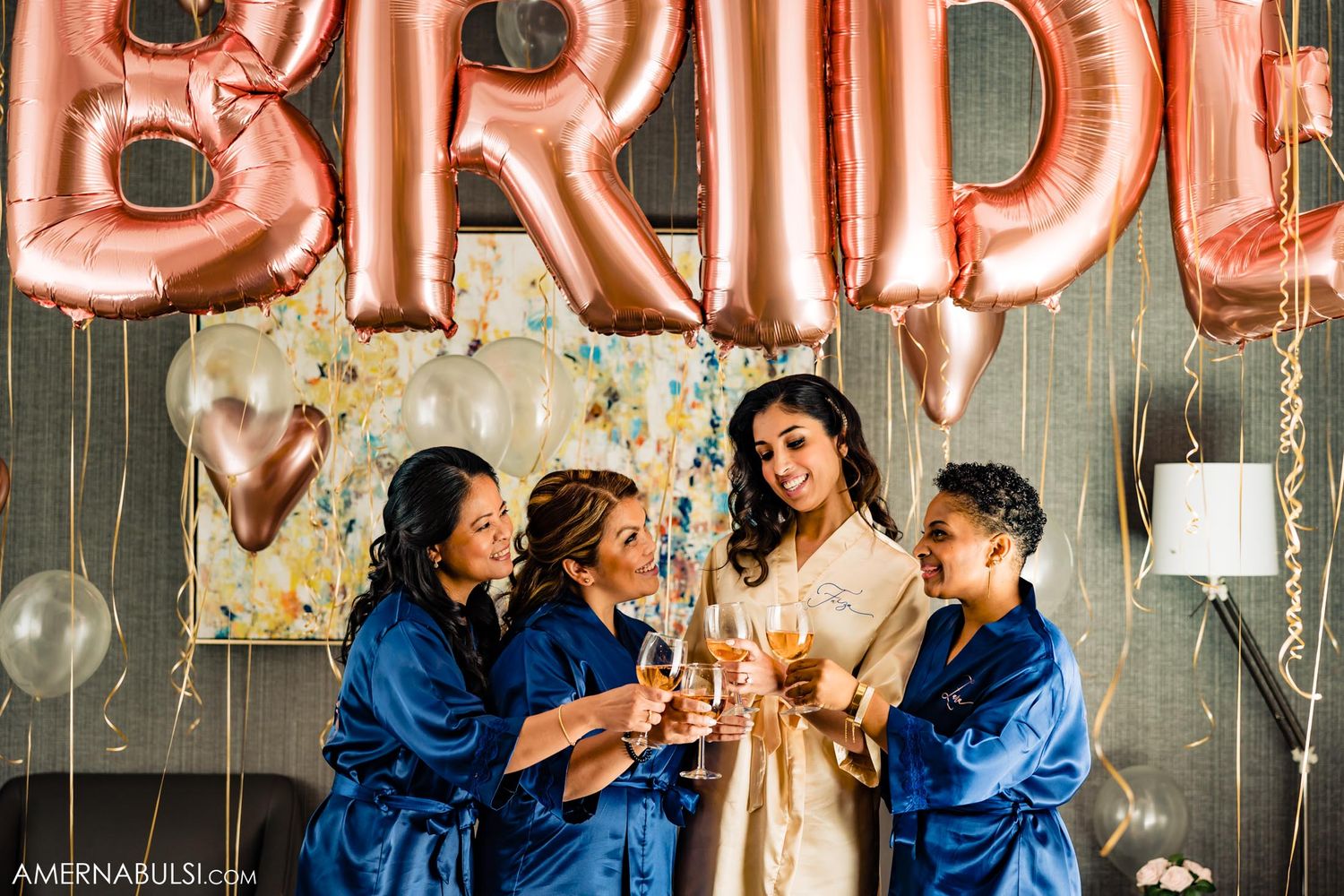 Wedding party in blue robes toasting with champagne under rose gold BRIDE balloons.