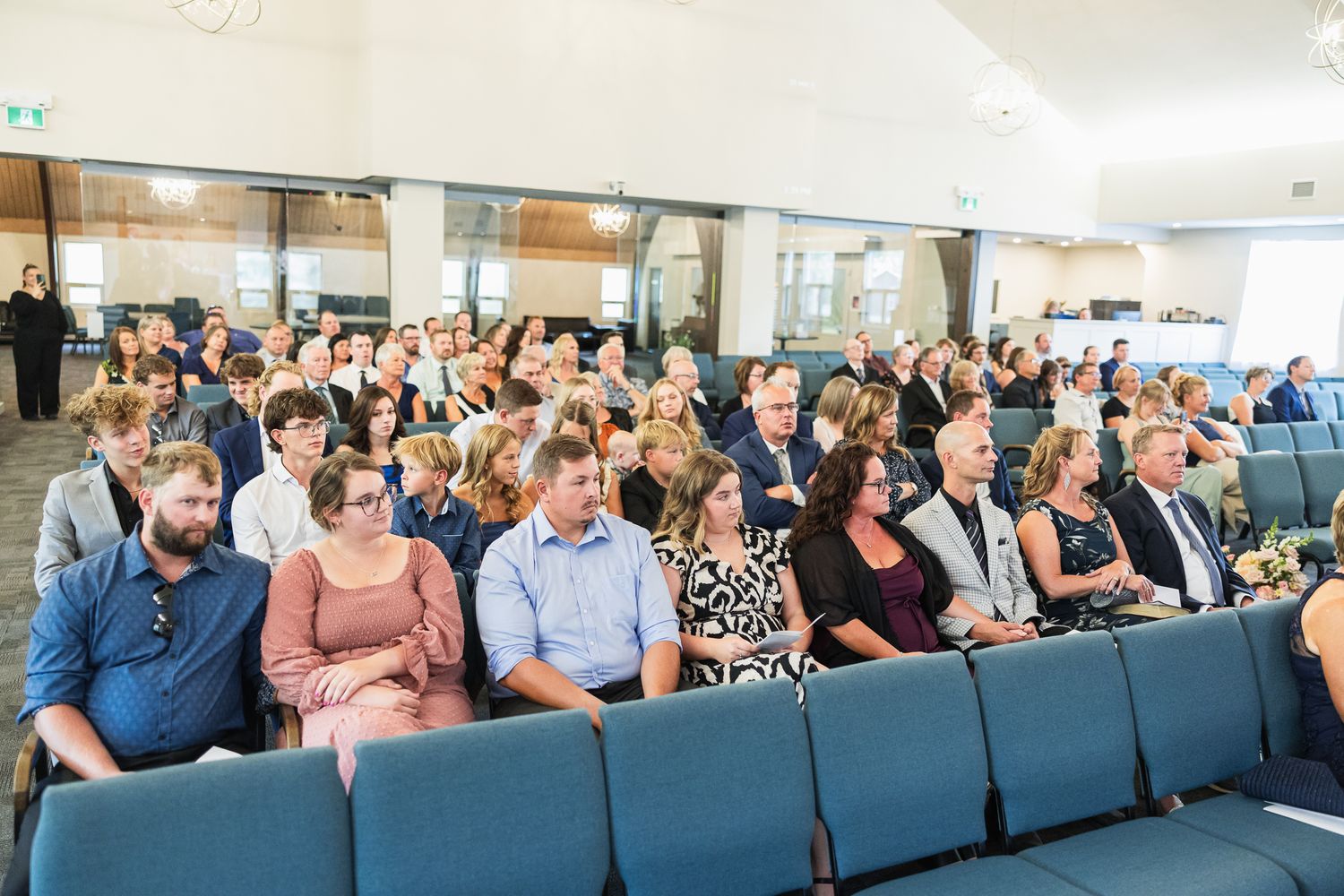 Wedding guests seated in rows of blue chairs at an indoor ceremony venue.
