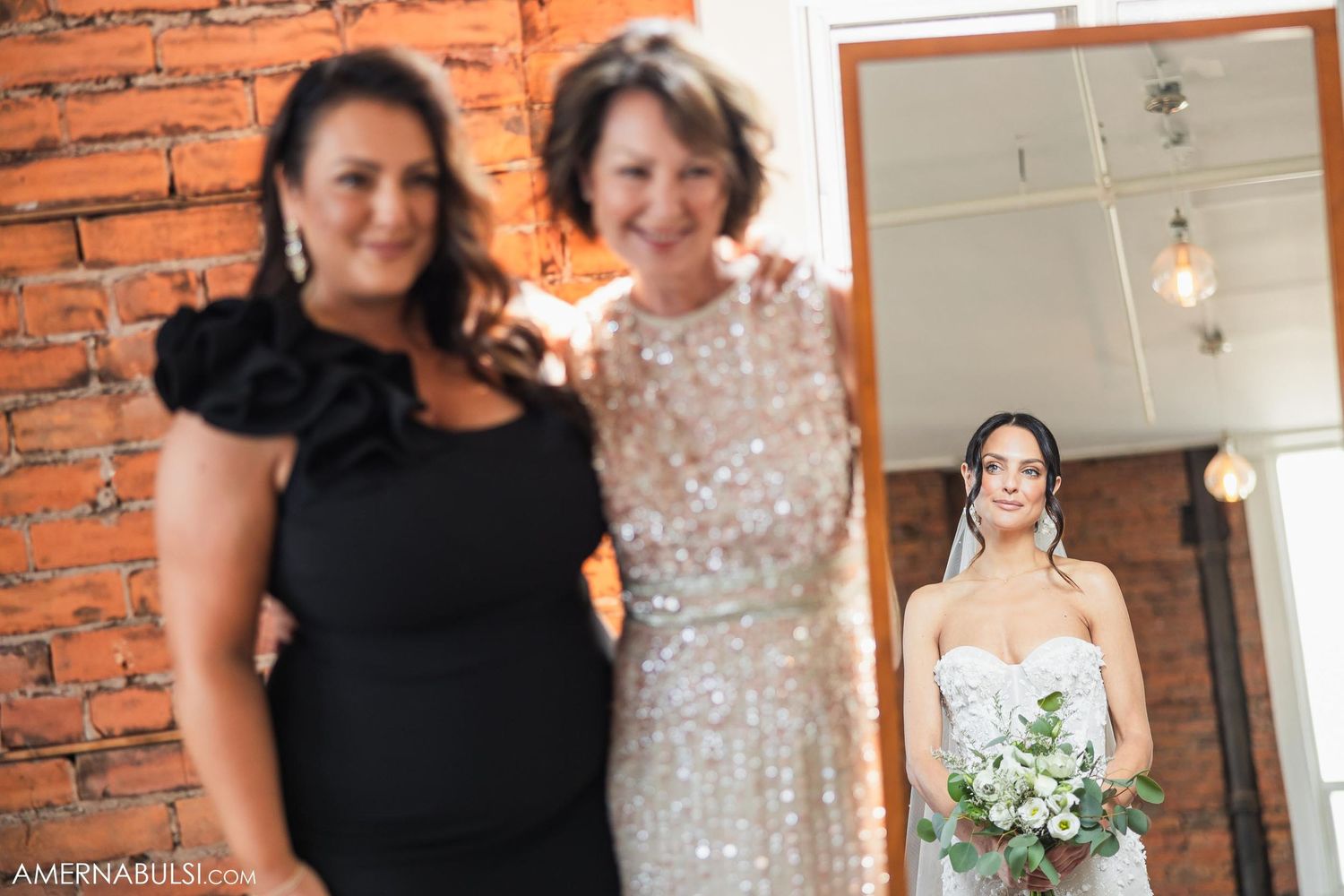 Three women pose together in front of brick walls during wedding preparations.