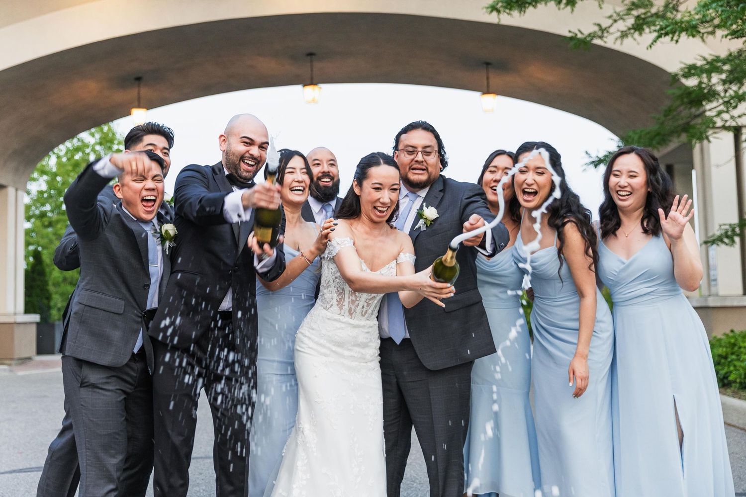 Bridal party poses together under an outdoor archway celebrating with champagne.