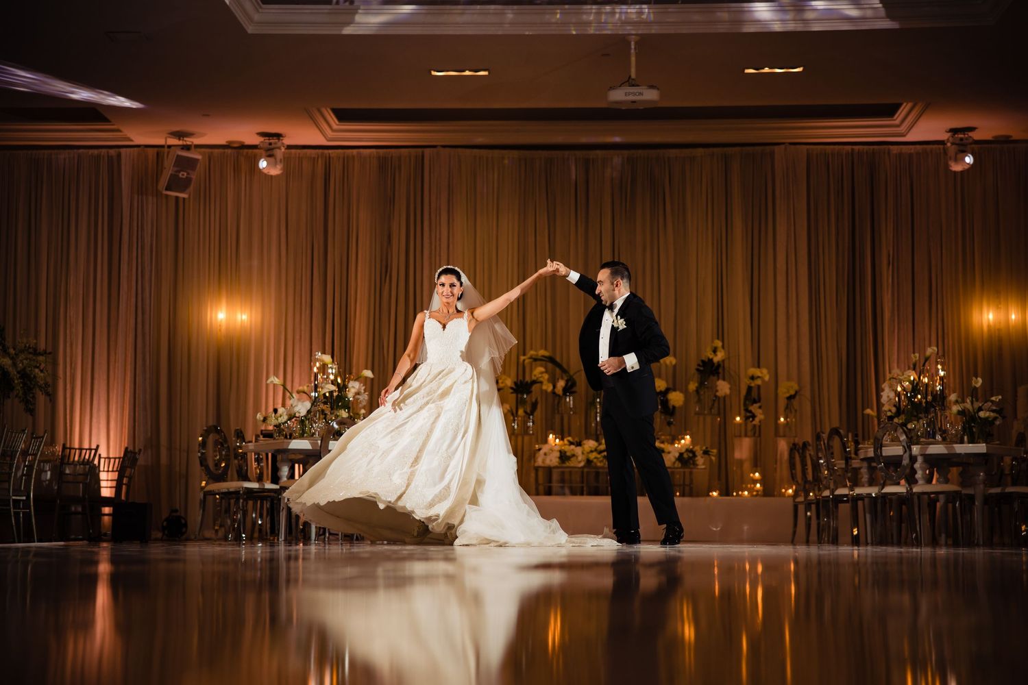 A romantic couple sharing their first dance in an elegant ballroom with warm lighting and curtained backdrop.