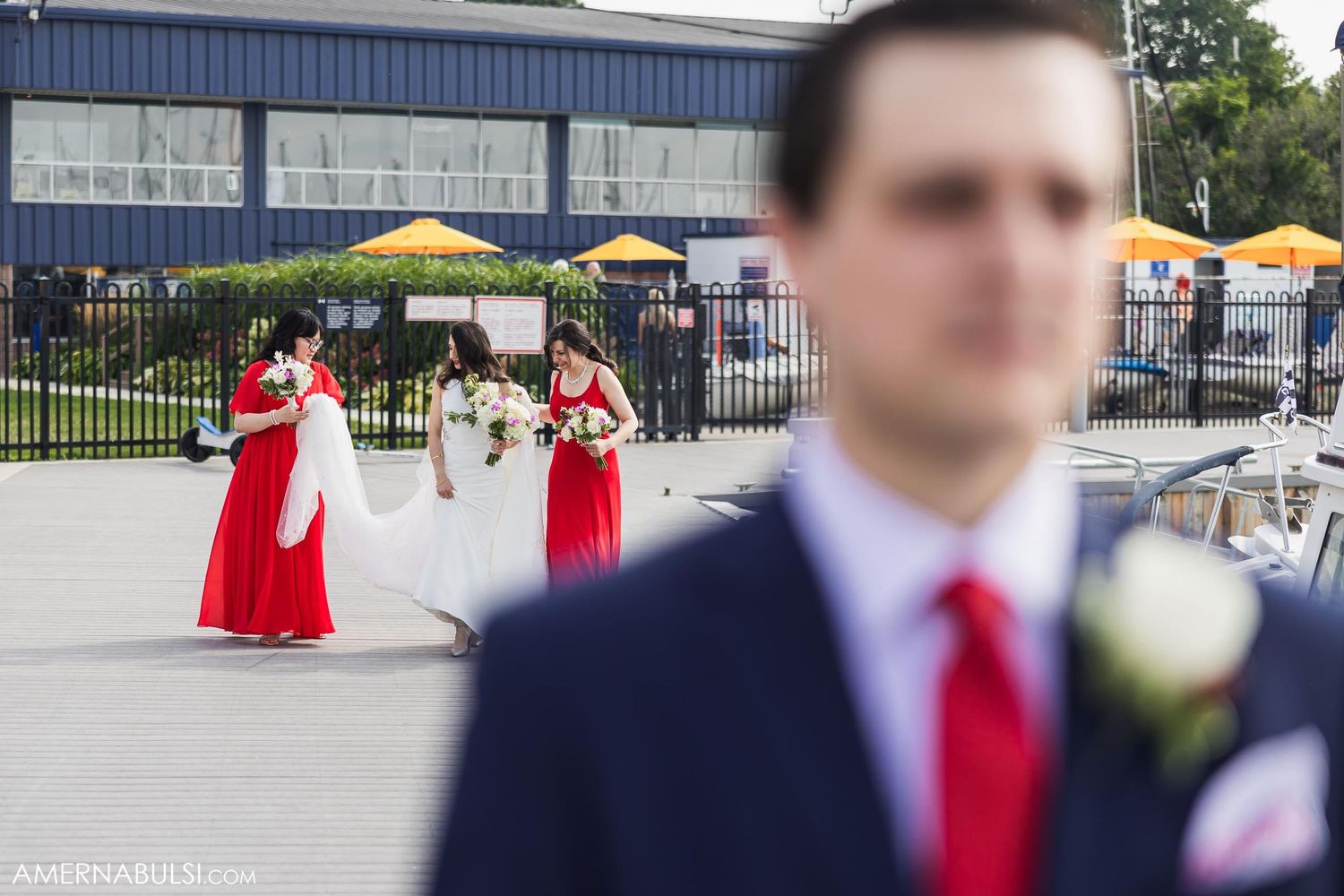 Wedding party members in red dresses gather outside venue while a suited figure stands in foreground.