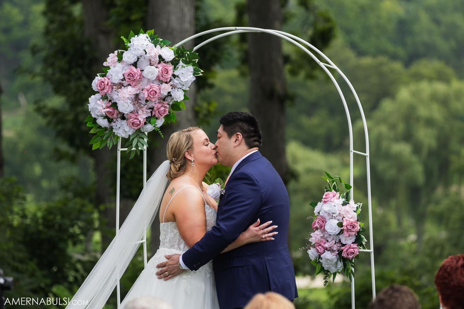 Couple shares first kiss under white arch decorated with pink and white floral arrangements.