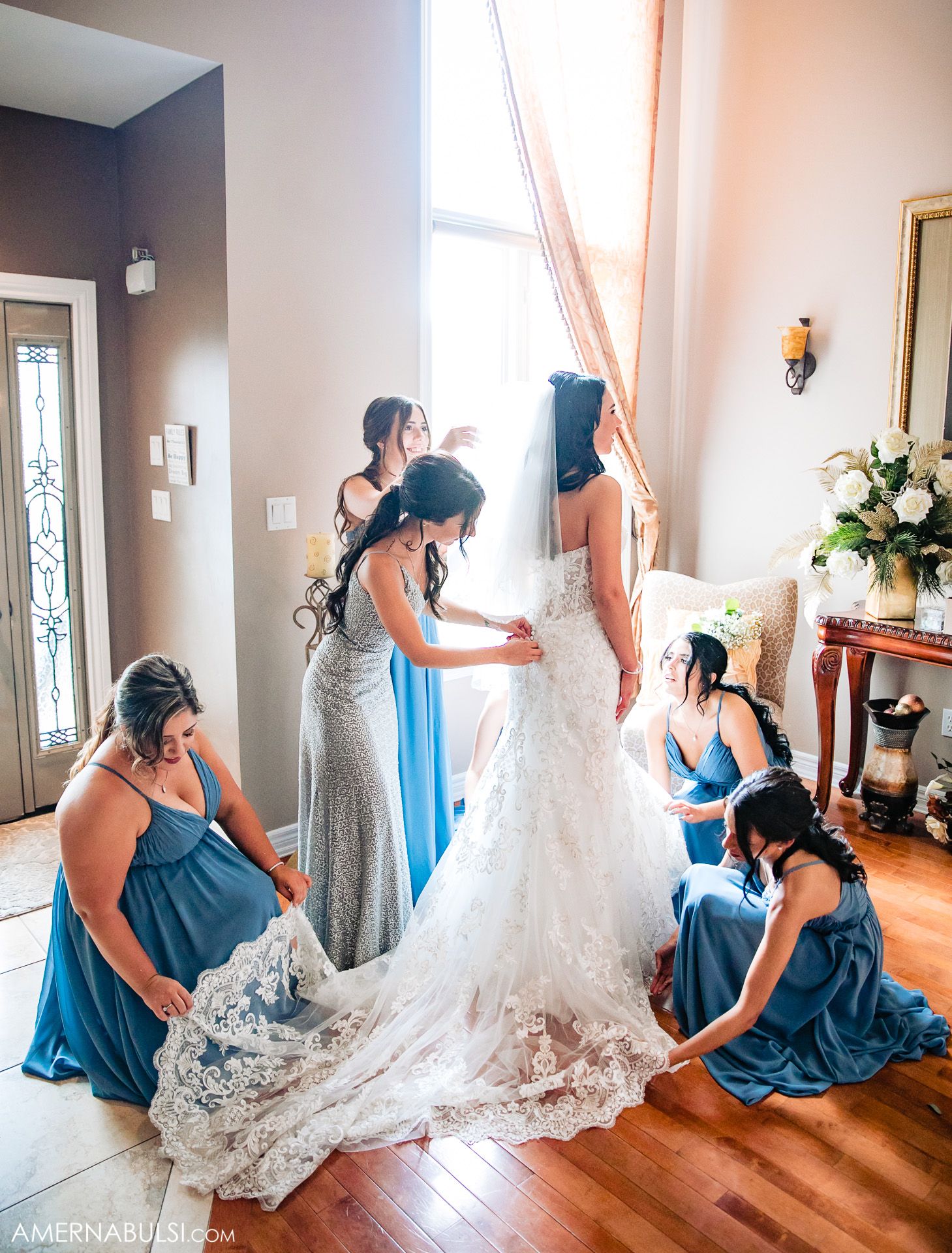 Bridesmaids in blue dresses help adjust the train of a lace wedding gown in an elegant room with hardwood floors.
