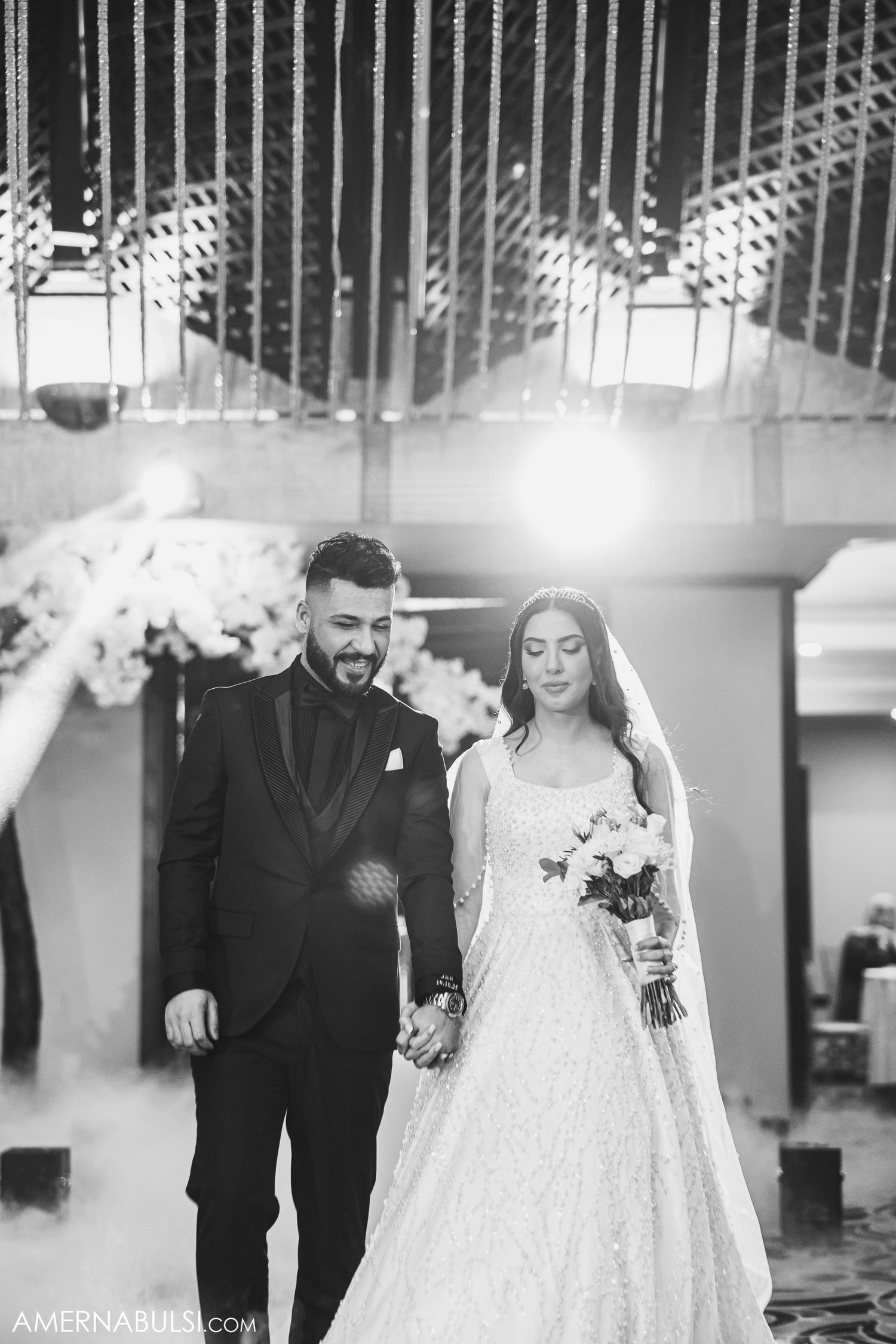 A black and white wedding photo showing a couple in formal attire walking together under decorative lighting.