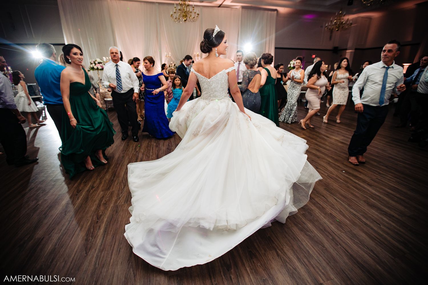 A bride in a flowing white gown twirls on the dance floor surrounded by wedding guests during the reception.