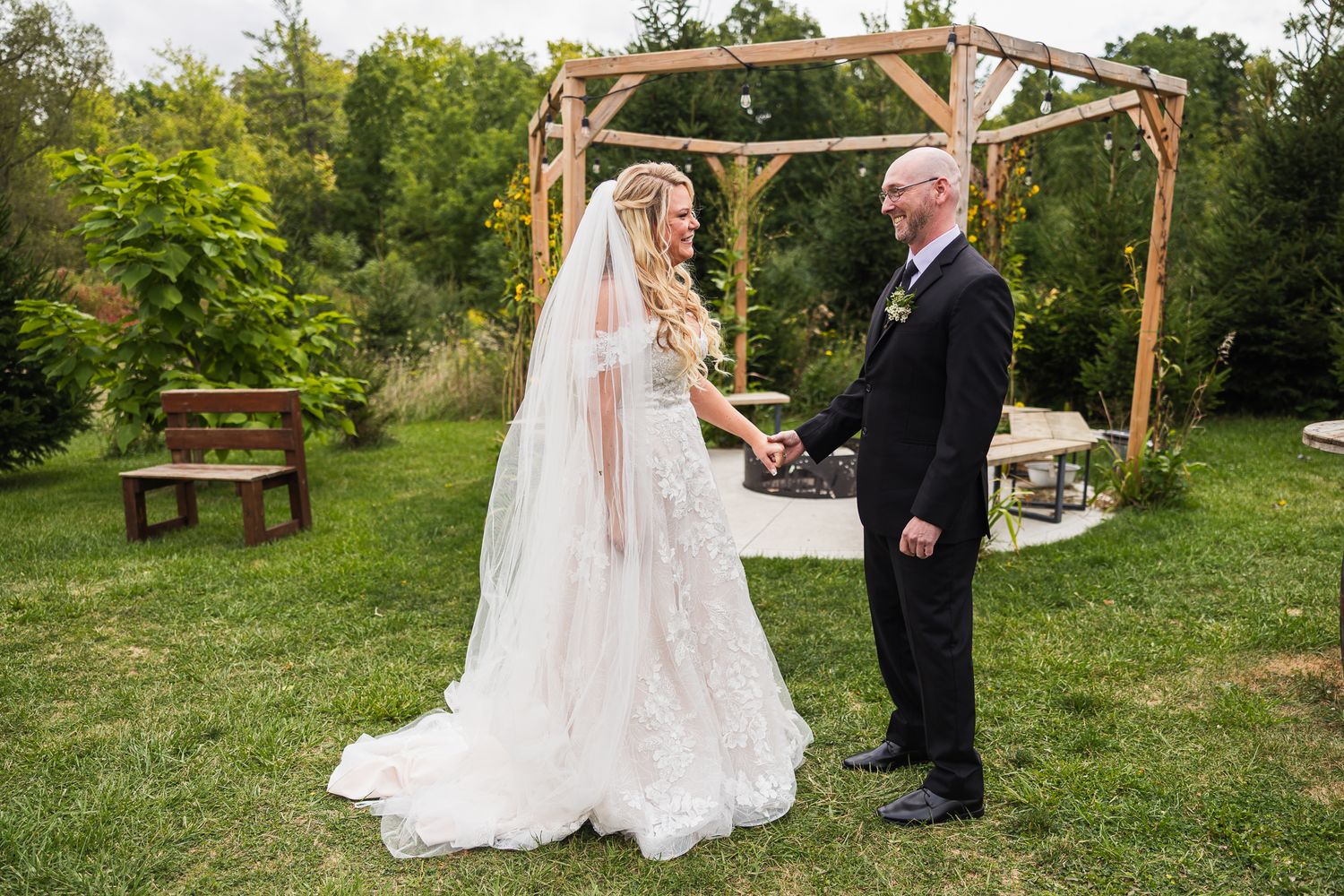 Wedding ceremony takes place under a rustic wooden arbor decorated with flowers in a scenic outdoor garden setting.