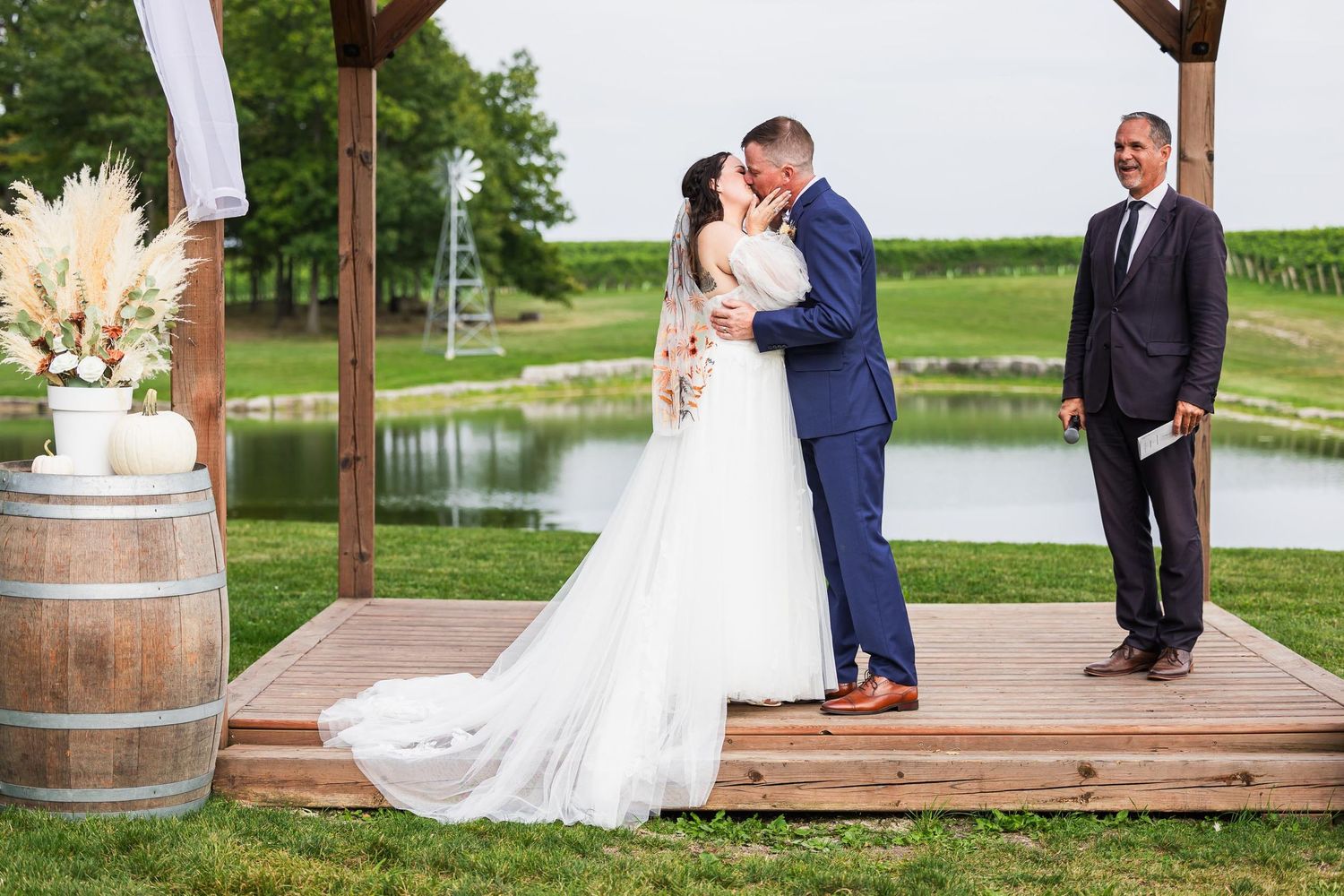 Romantic wedding moment on wooden deck with wine barrel decor and pond backdrop.