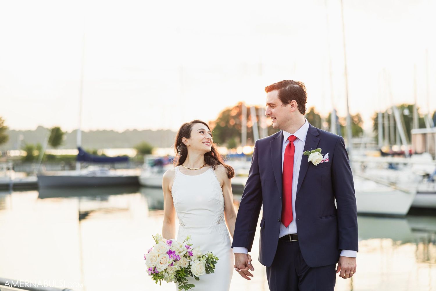 Newlyweds walk together by a marina holding a purple and white bouquet.
