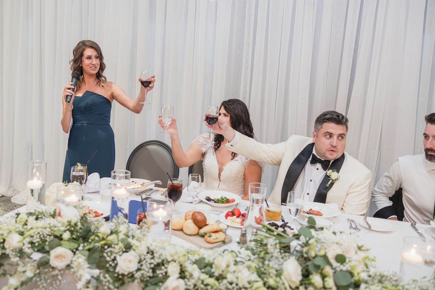 Wedding toast at reception table with guests raising wine glasses, floral centerpieces, and woman in blue dress speaking.