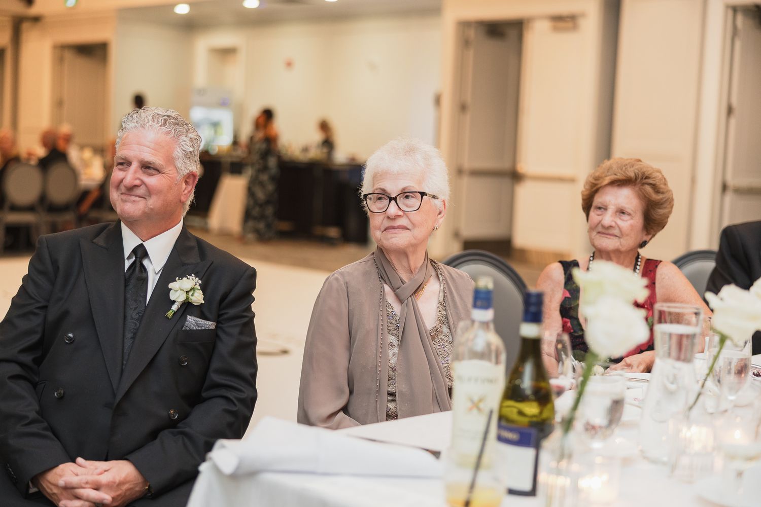 Three elderly guests seated at a wedding reception table, smiling and watching the ceremony proceedings.