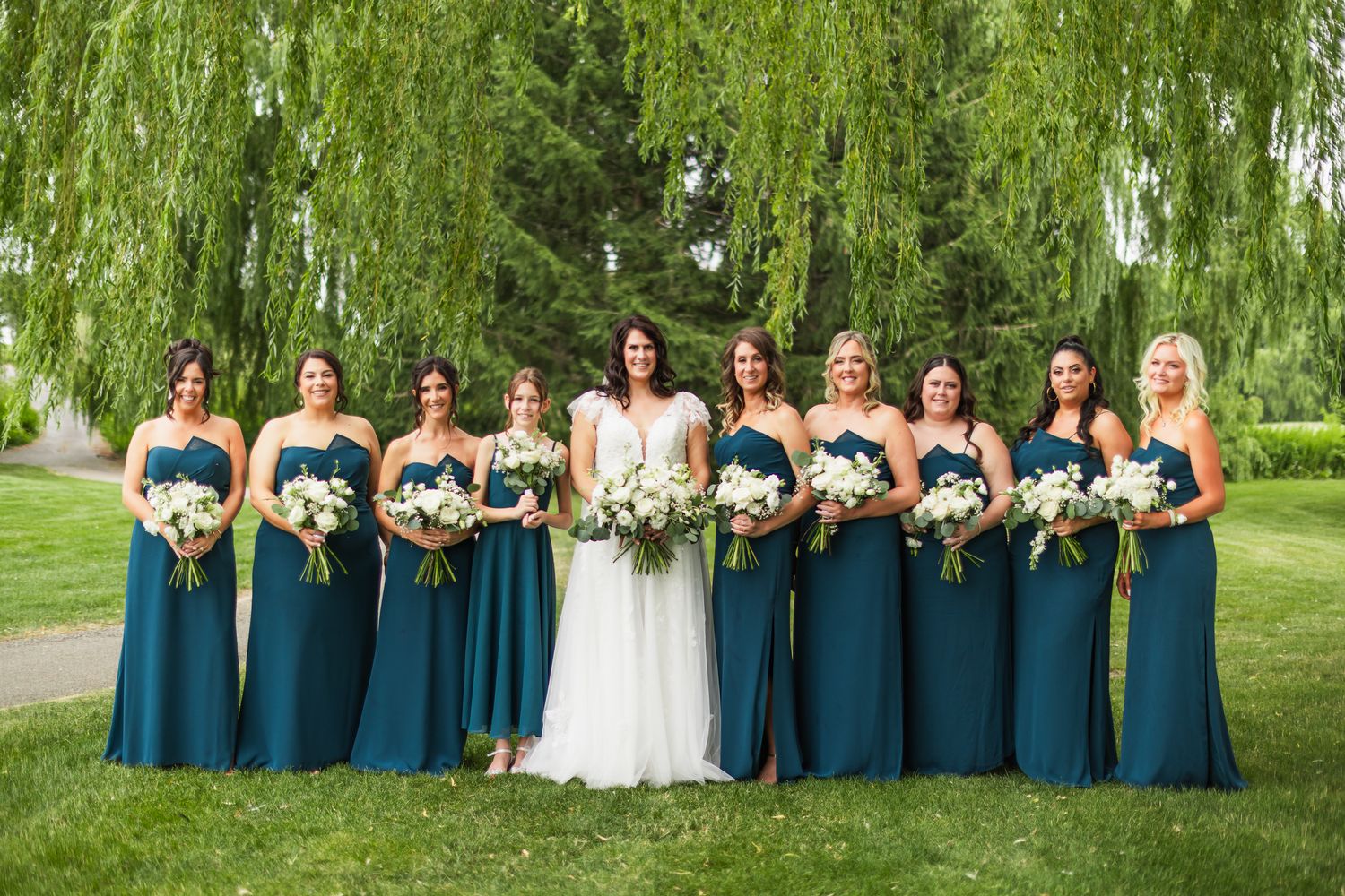 Bride poses with eight bridesmaids in teal gowns holding white bouquets under a weeping willow tree.
