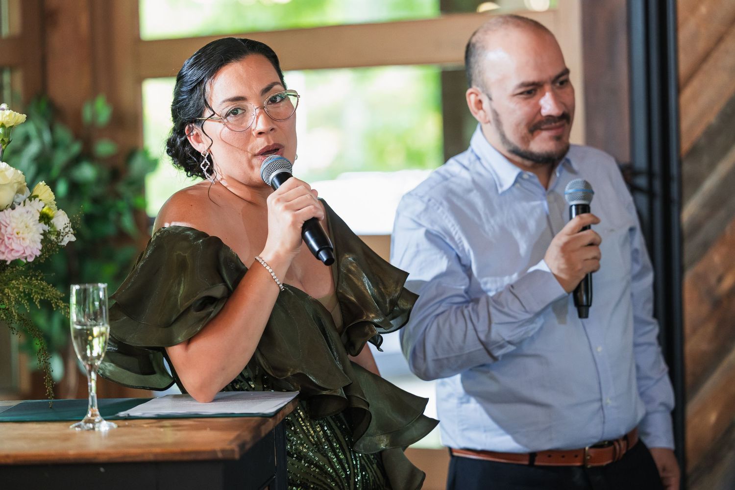 A woman in an olive dress speaks into a microphone at an event, while a man in a blue shirt holds a mic beside her.