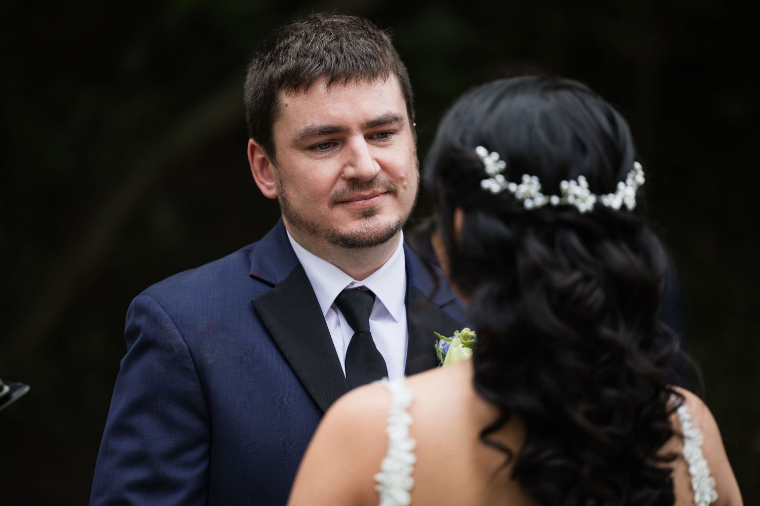 Groom in navy suit smiles at bride with floral hair crown during outdoor wedding ceremony.