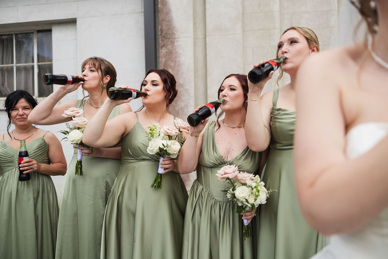 Bridesmaids in sage green dresses drink from Coca-Cola bottles while holding bouquets before wedding ceremony.