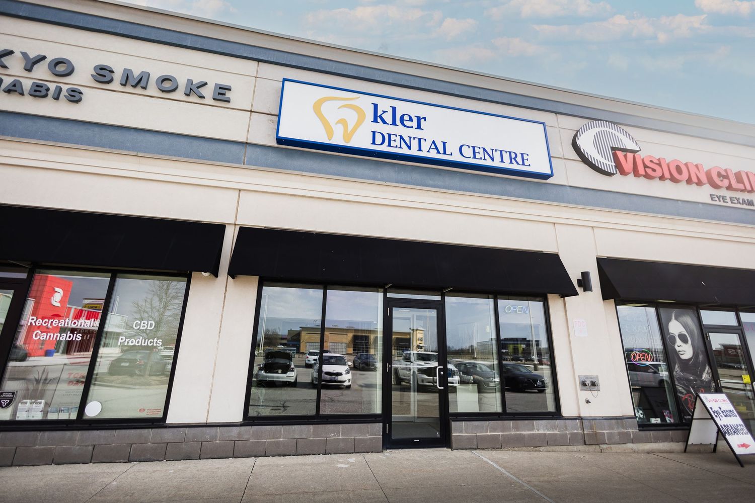 Exterior view of Kler Dental Centre in a strip mall, featuring large windows and signage in a suburban shopping plaza.