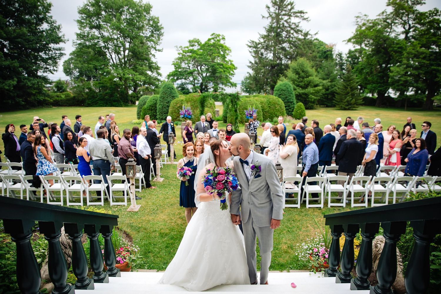 Outdoor garden wedding ceremony with guests seated on white chairs amid lush greenery.