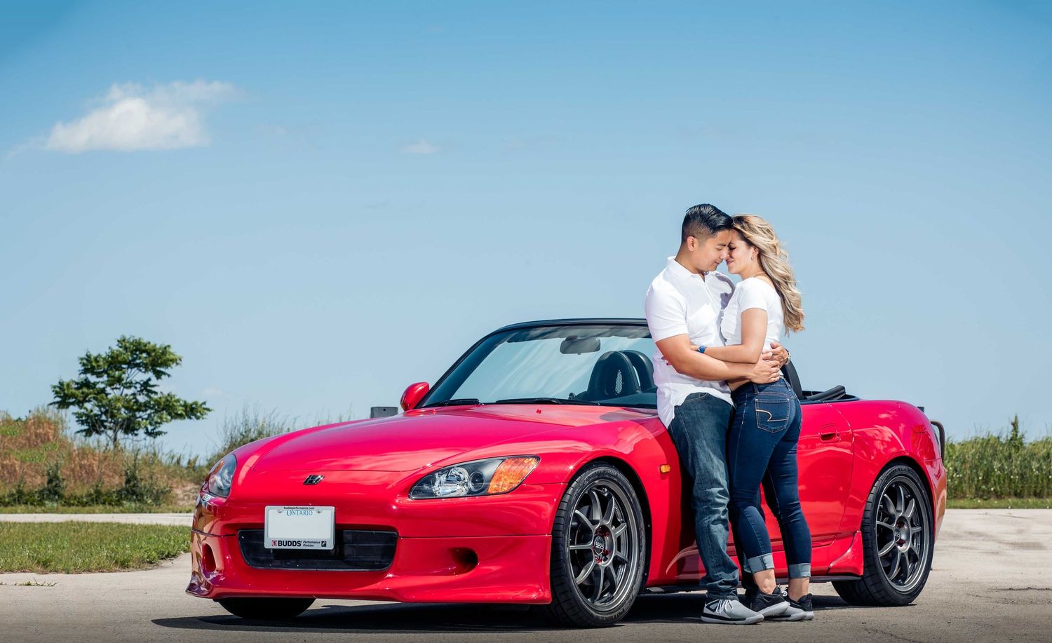 Series of romantic poses with a red sports car on an empty road against blue sky.