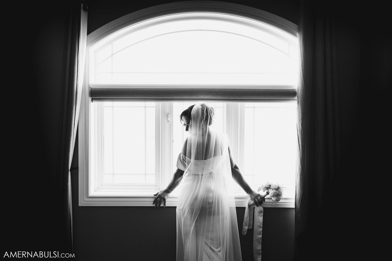 Silhouette of bride in wedding dress standing by window in dramatic lighting.