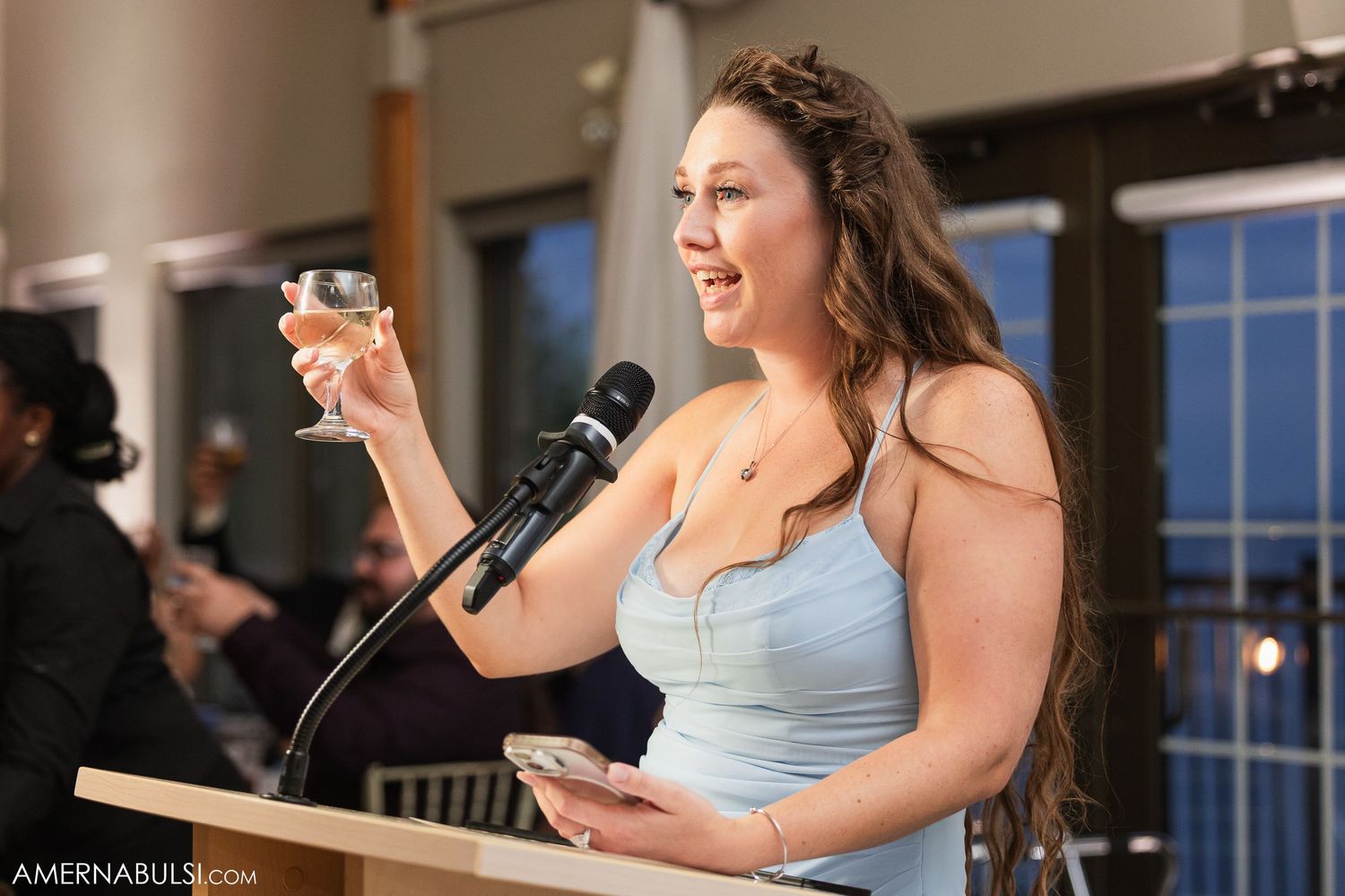 Person in light blue dress gives speech at formal event with ocean view through large windows.