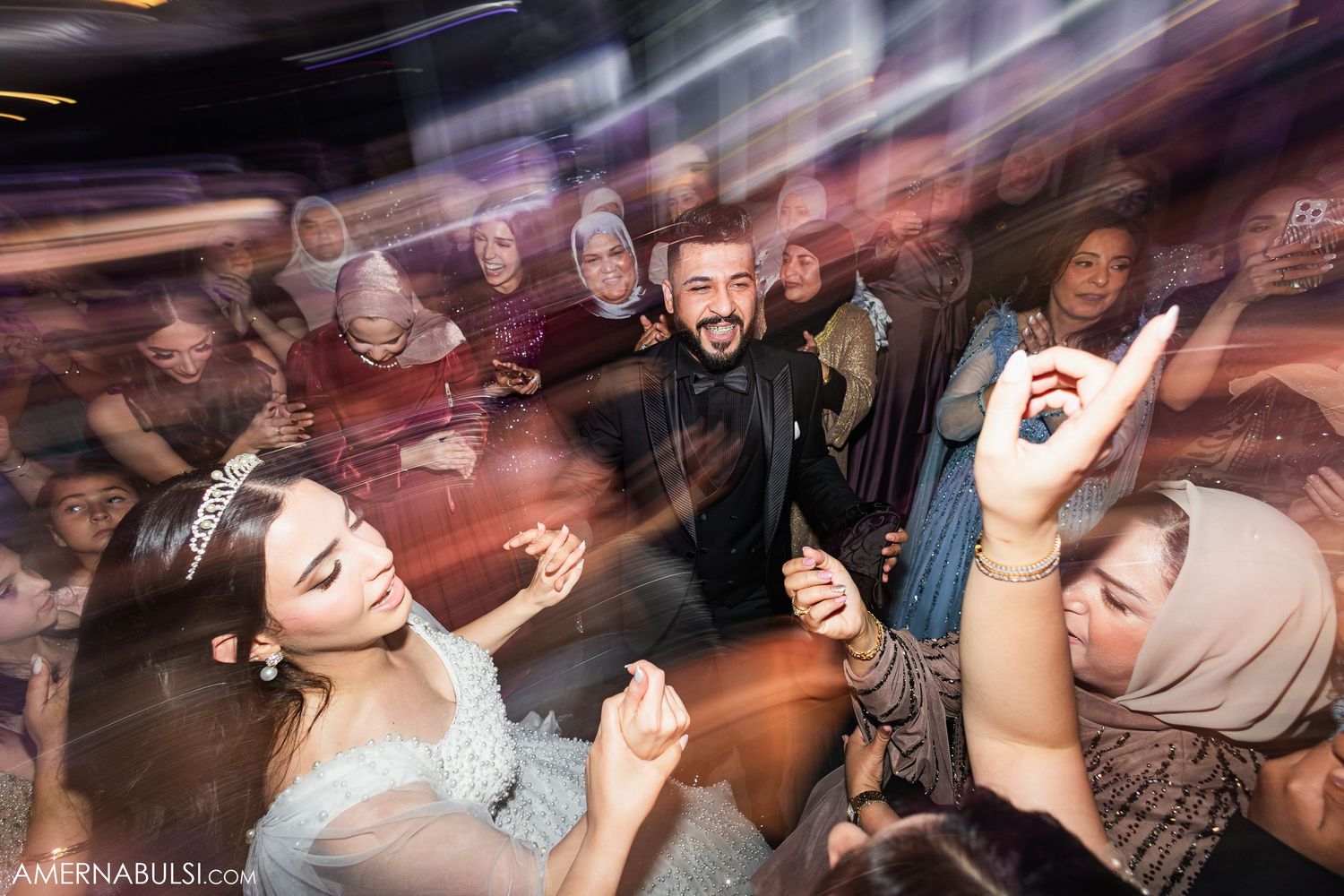 Energetic crowd dances and celebrates at a lively wedding reception with purple lighting effects.