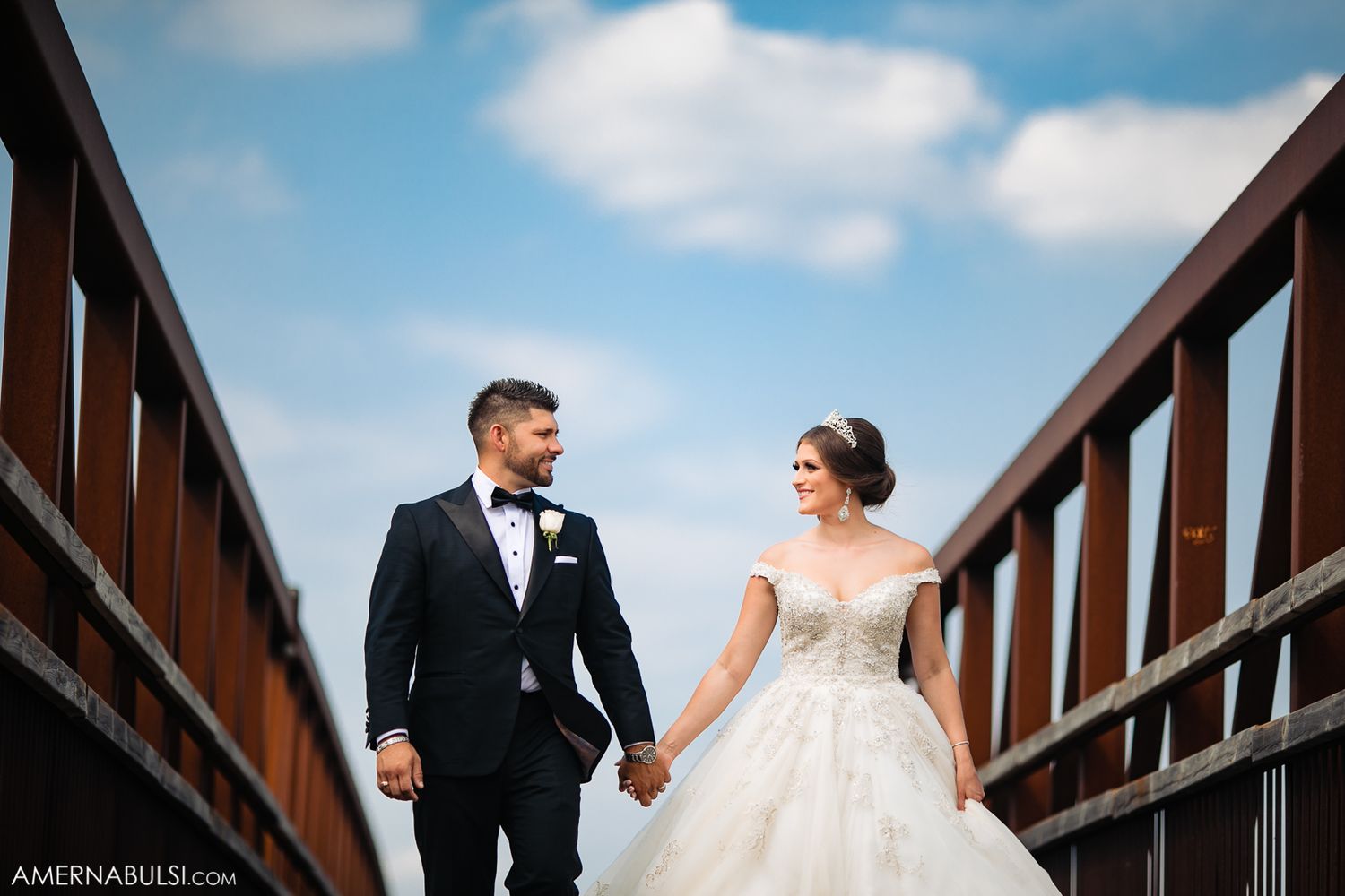 Elegant wedding couple poses on a rustic wooden bridge against a blue sky backdrop.