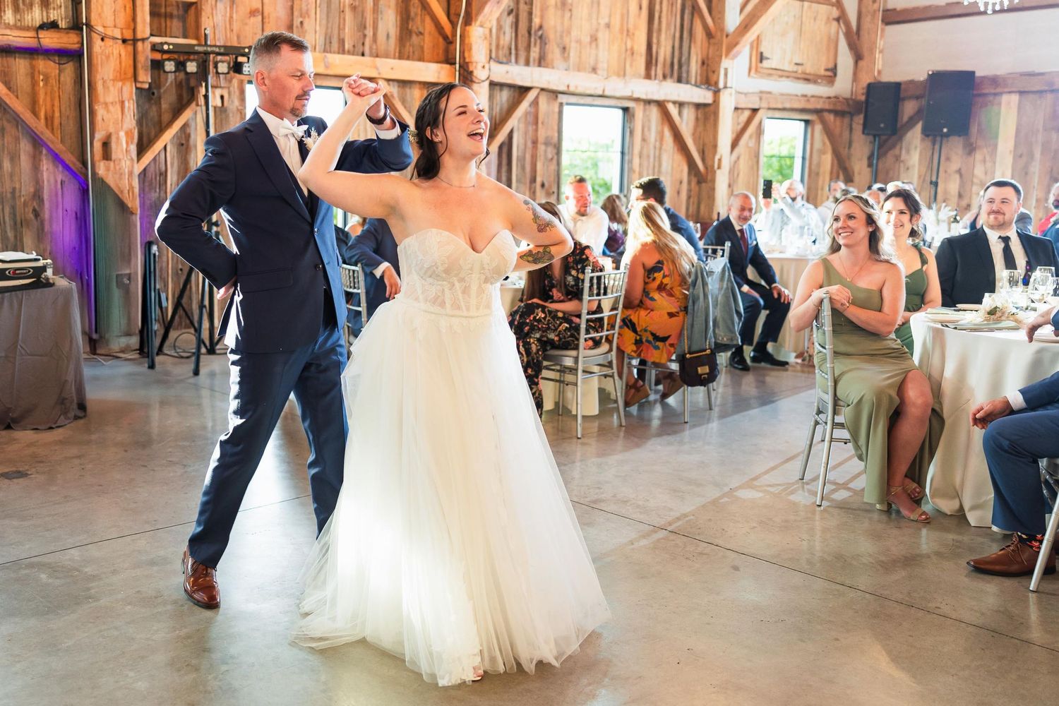 Wedding couple dances at rustic barn reception while guests look on and applaud.