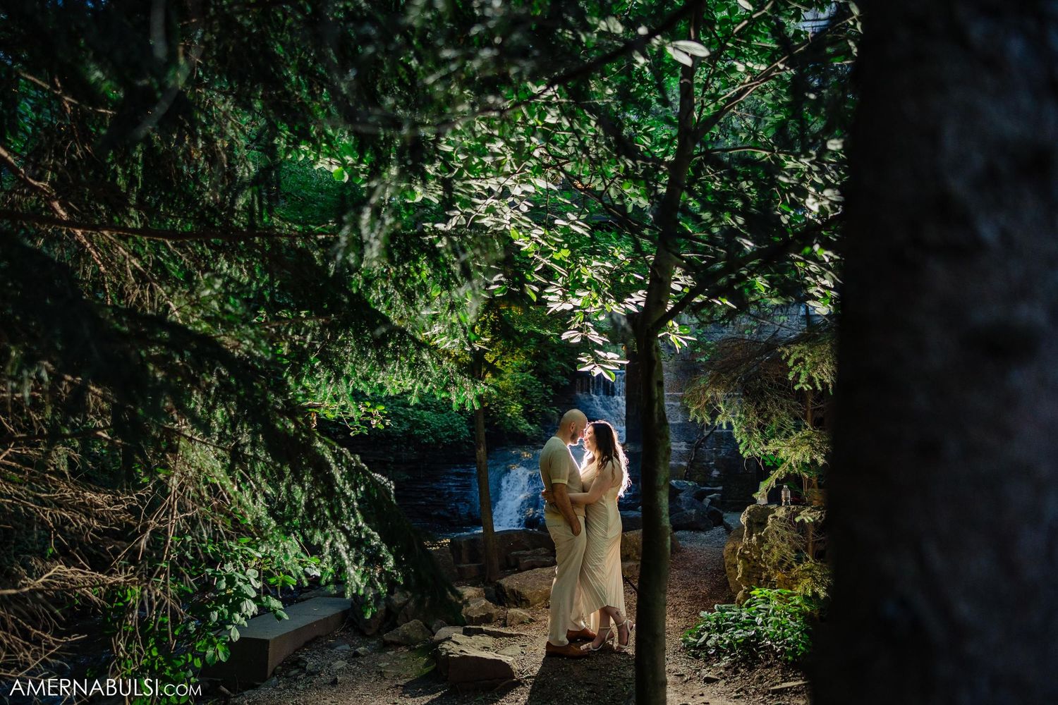 Couple in neutral attire walking along a scenic woodland path near a waterfall.