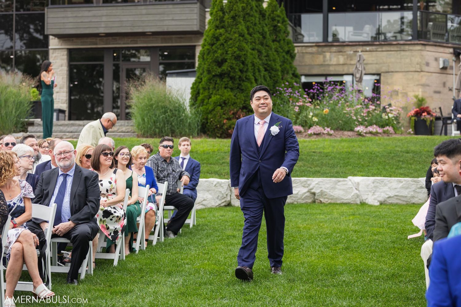 Groom walks down outdoor ceremony aisle with guests seated on white chairs.
