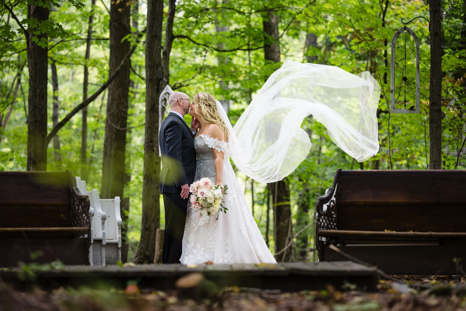 Bride's veil billowing dramatically in wind during outdoor forest wedding photoshoot.