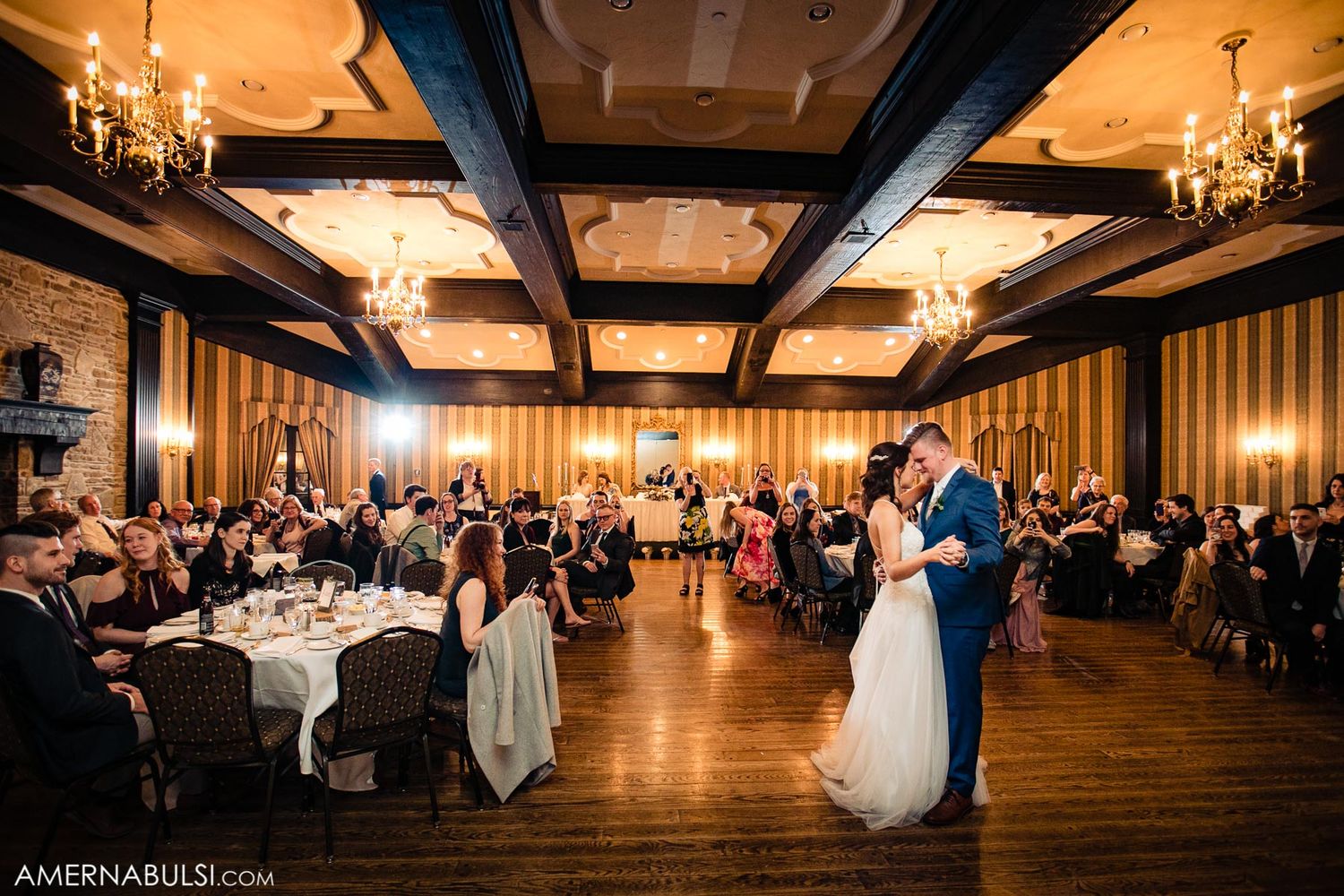 Panoramic view of an elegant wedding reception hall with wooden beams, chandeliers and guests seated at round tables.