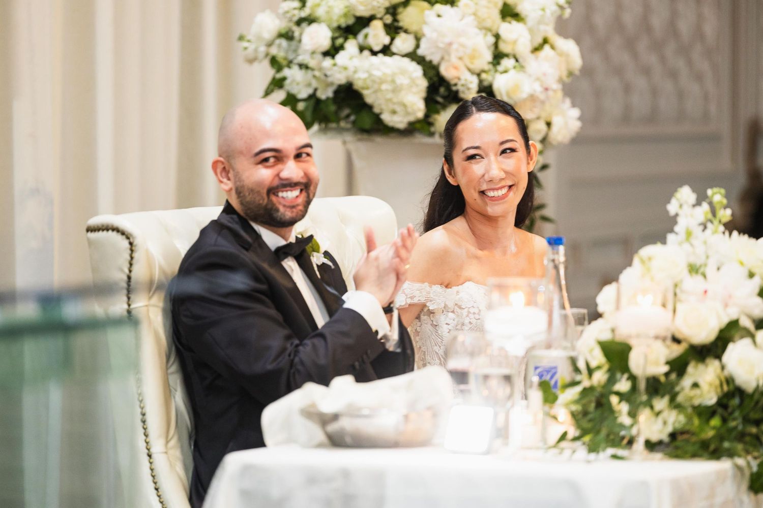 Wedding couple shares laughter at reception table decorated with white flowers.