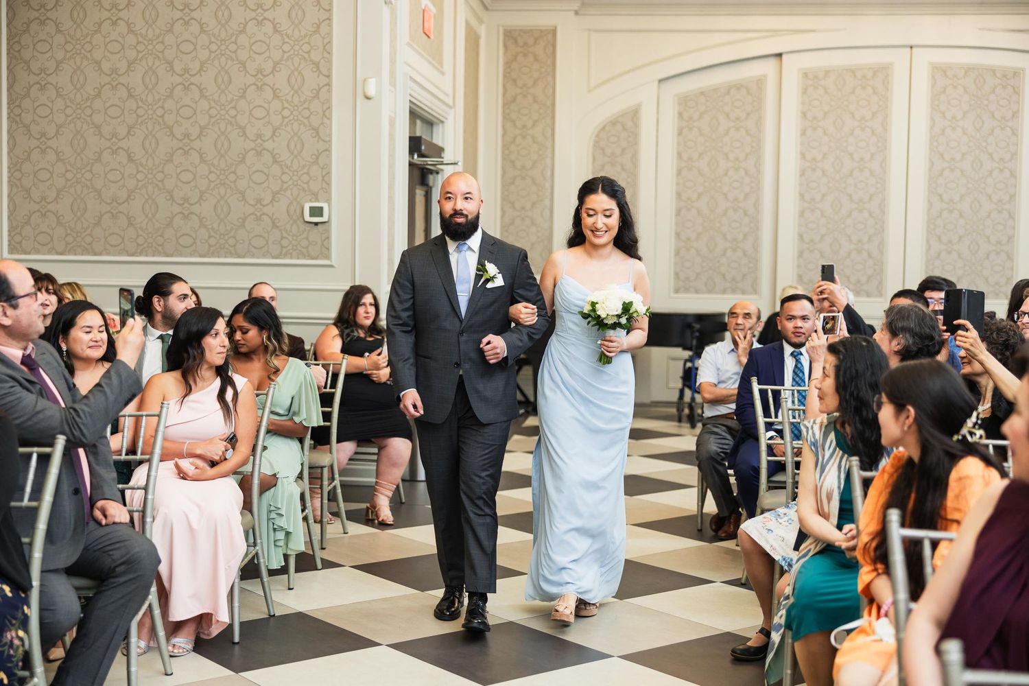 Wedding party processes down aisle while guests capture moment on phones in formal ballroom.