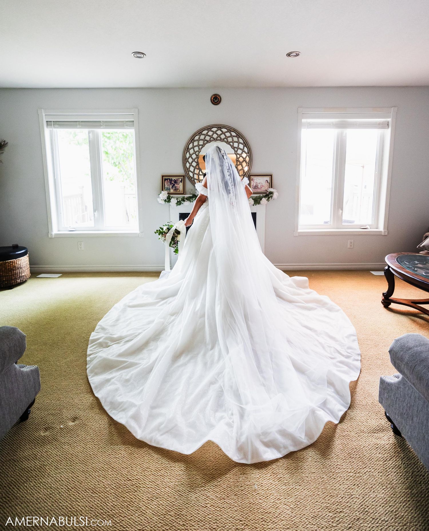 Back view of bride in flowing white wedding gown with long train standing in sunlit room.