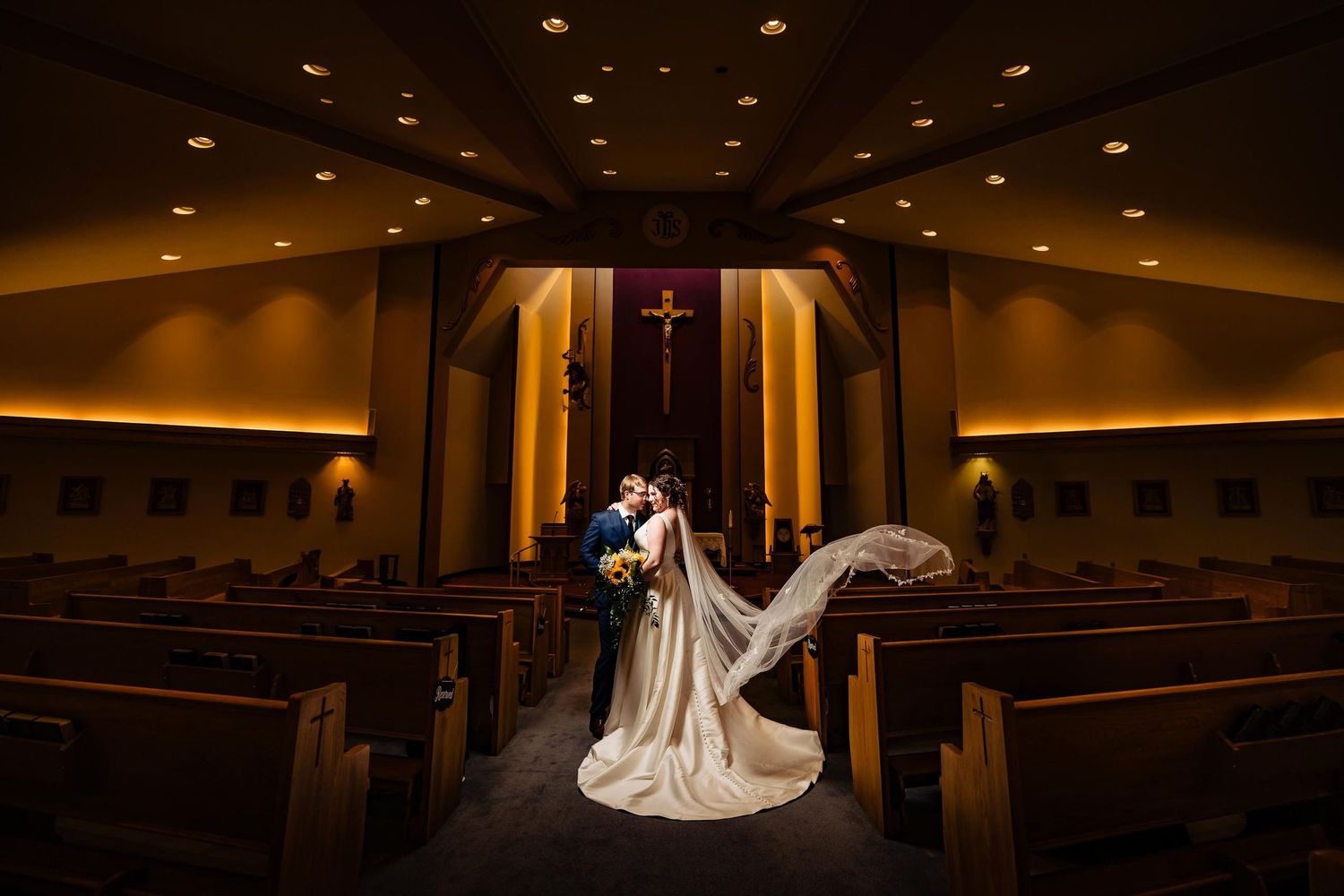 Dramatic church wedding photo with flowing veil and illuminated cross in background.