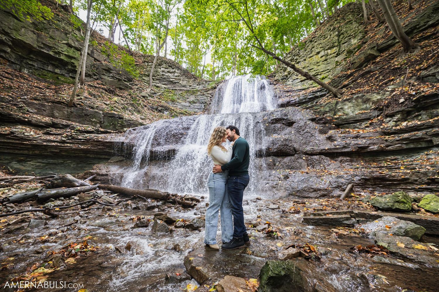 A couple embraces in front of a scenic waterfall cascading down rocky cliffs.