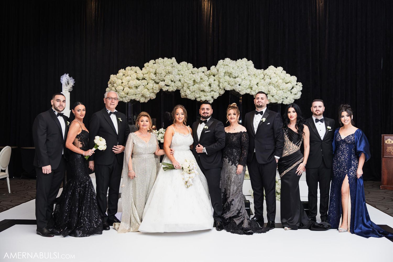 Wedding party in formal attire posing against backdrop with white floral arrangements.