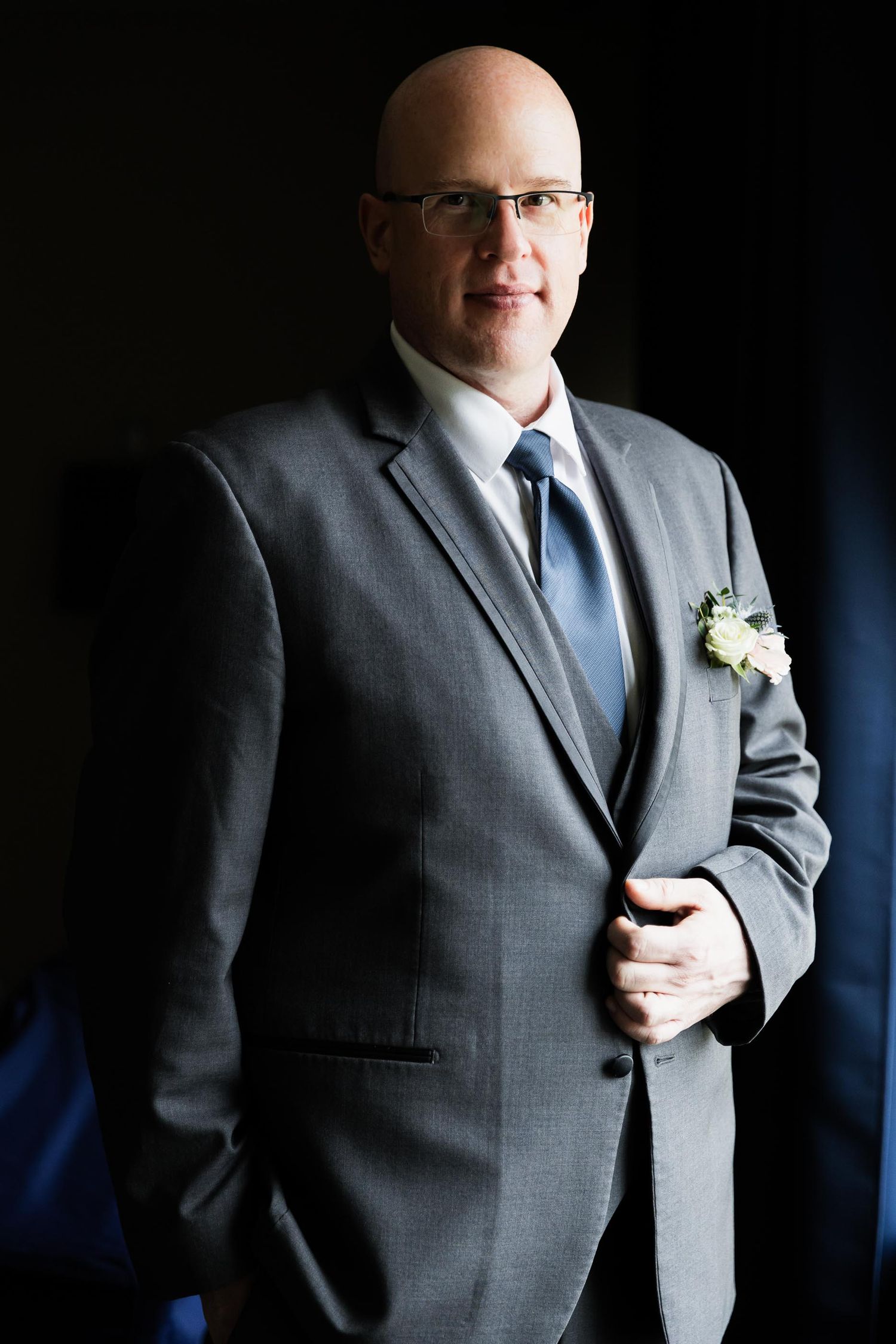A formal portrait of a person in a grey suit with a boutonniere against a dark background.