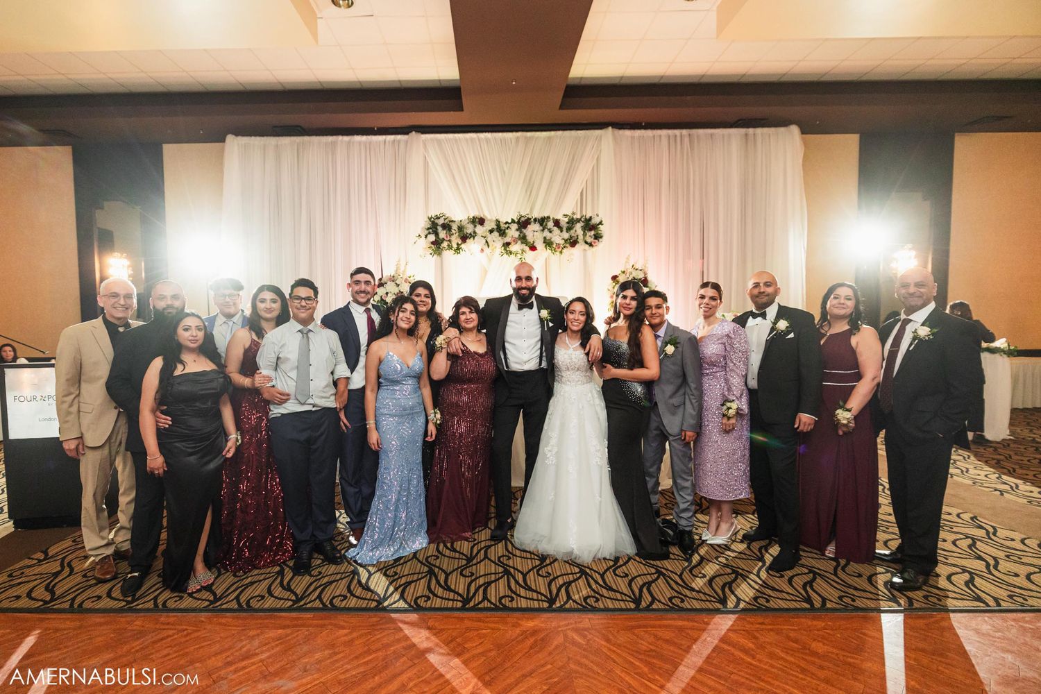 A large wedding party poses together for a formal group photo in front of a decorated backdrop.