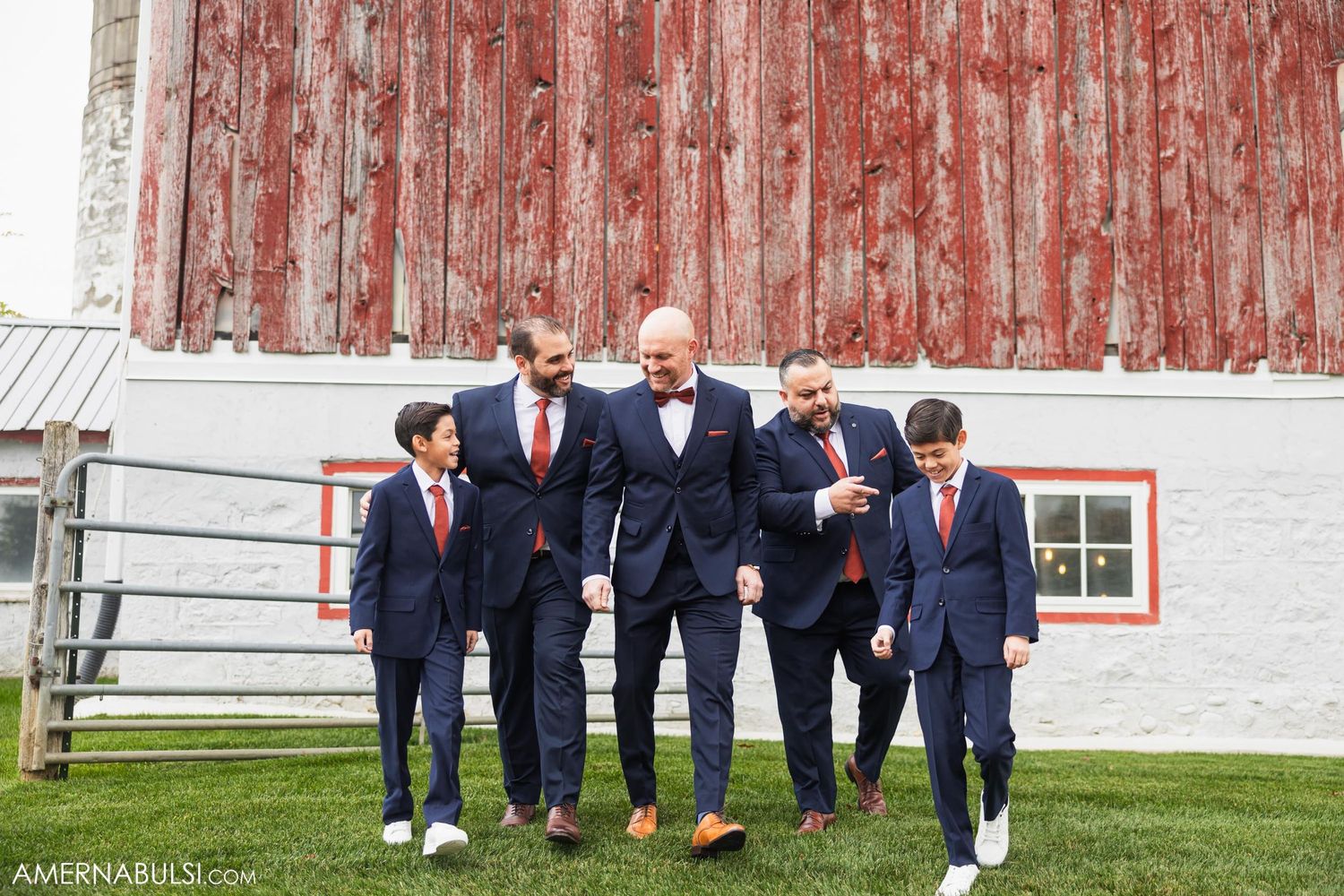 Wedding party in coordinated navy suits and red ties pose casually against red barn backdrop.