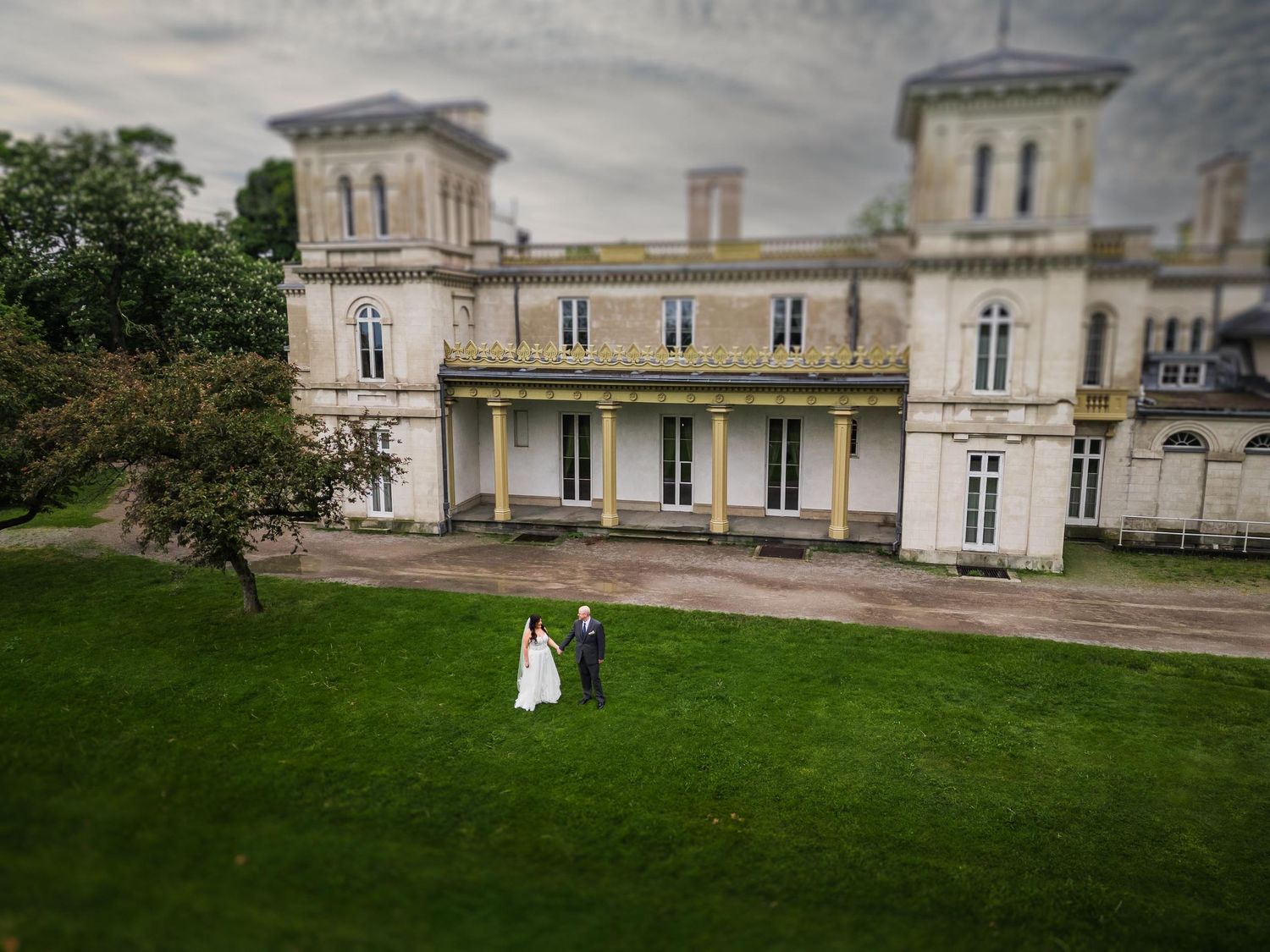 Aerial view of wedding couple standing on lawn in front of grand historic mansion.