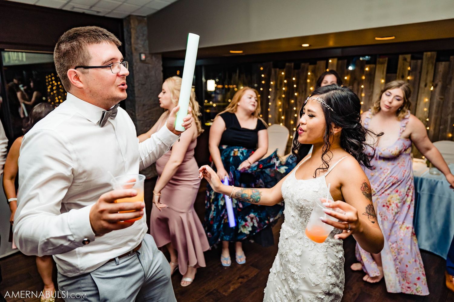 Party guests dancing with drinks and glowsticks at indoor celebration venue.