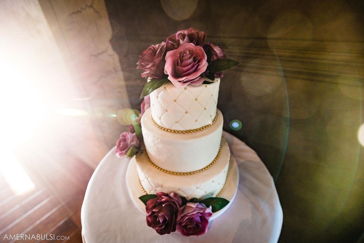 An elegant three-tiered white wedding cake decorated with deep burgundy roses and golden accents.