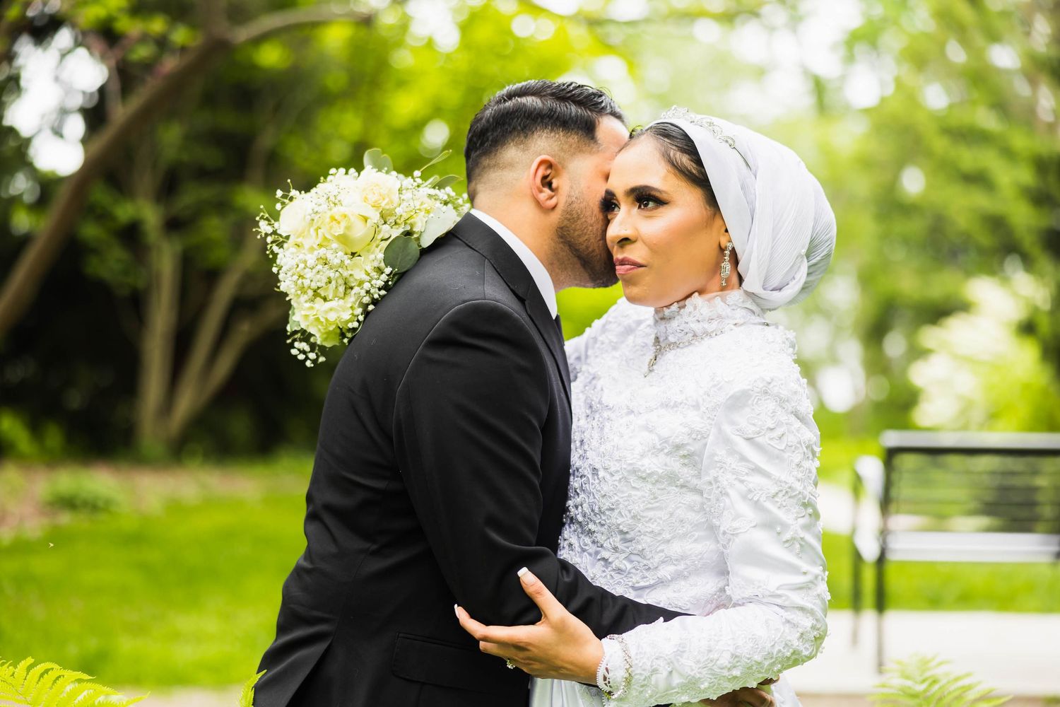 A couple dressed in wedding attire embrace in a lush garden setting with a white bouquet.