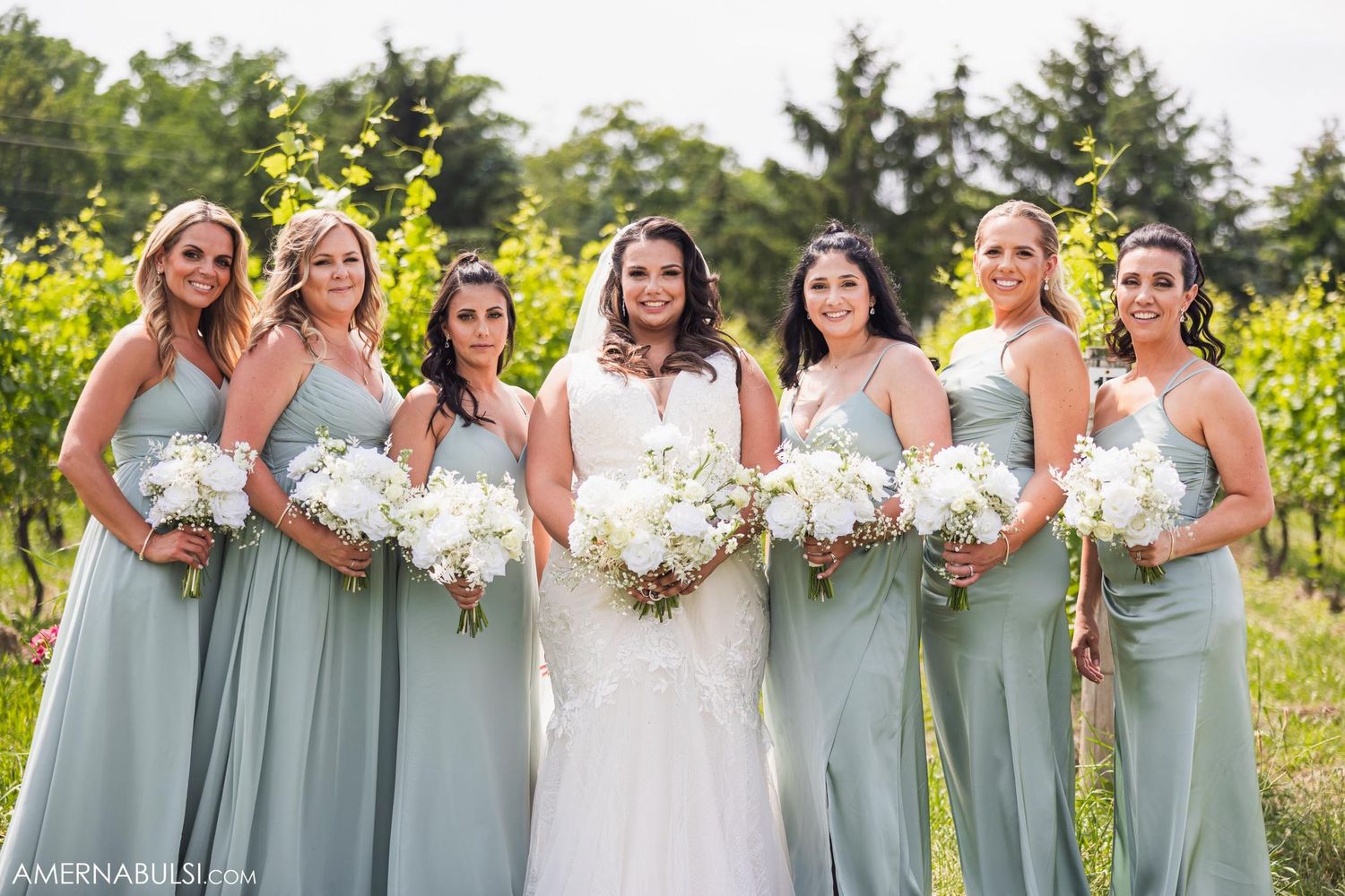 Formal portrait of bride and bridesmaids in sage green dresses holding bouquets in vineyard setting.