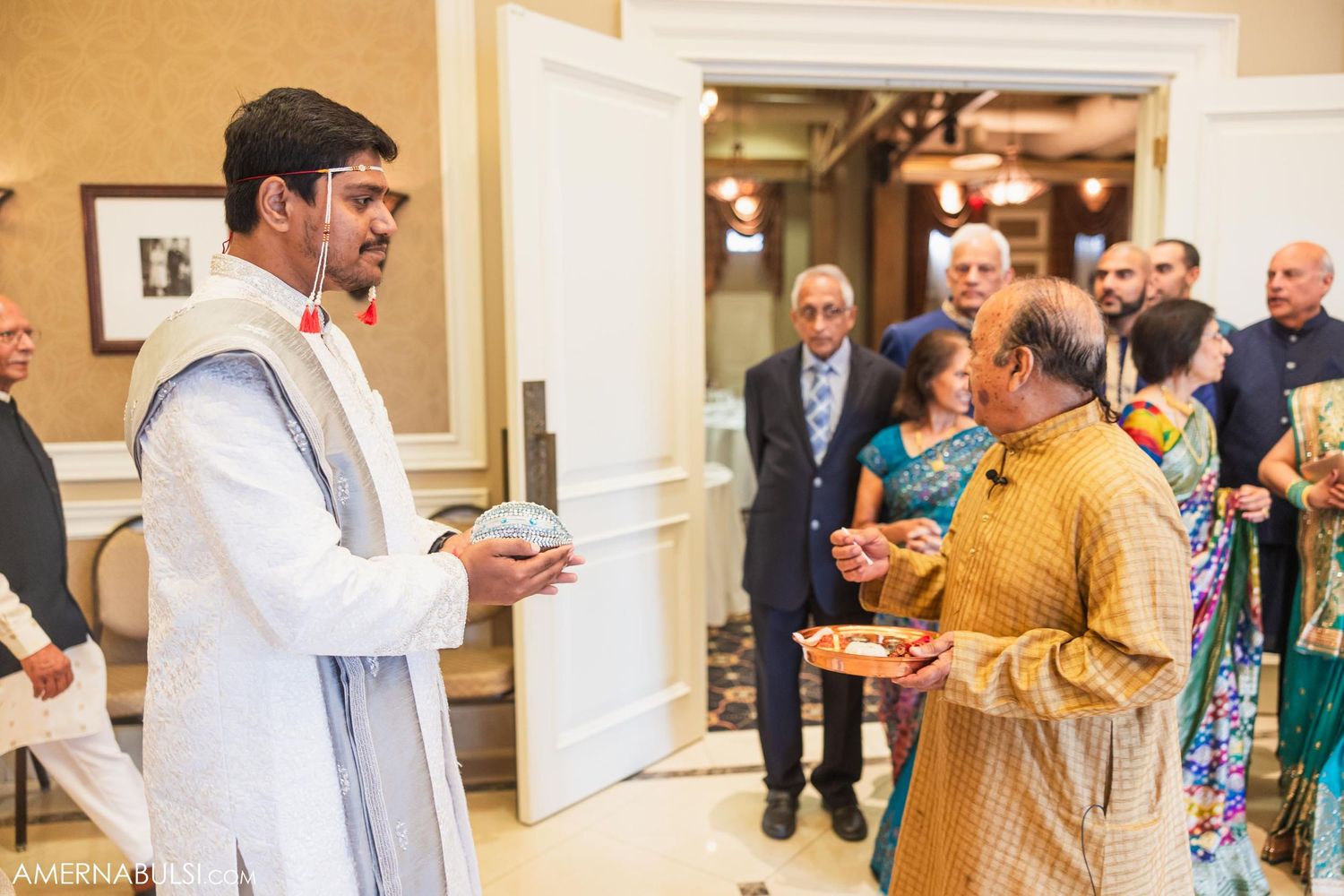 Two people greet each other in formal attire at an indoor gathering while others look on.
