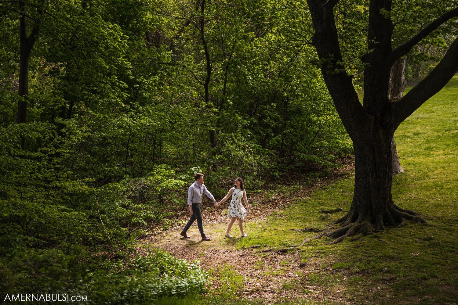 A couple walks hand in hand along a sunlit forest path beneath a large tree.