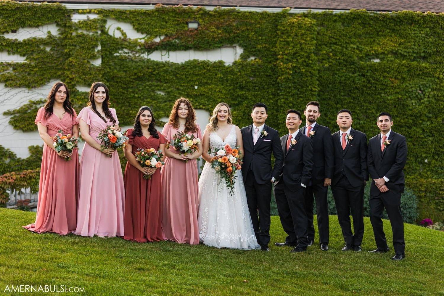 Wedding party in pink bridesmaid dresses and black suits poses on grassy hill for formal photo.