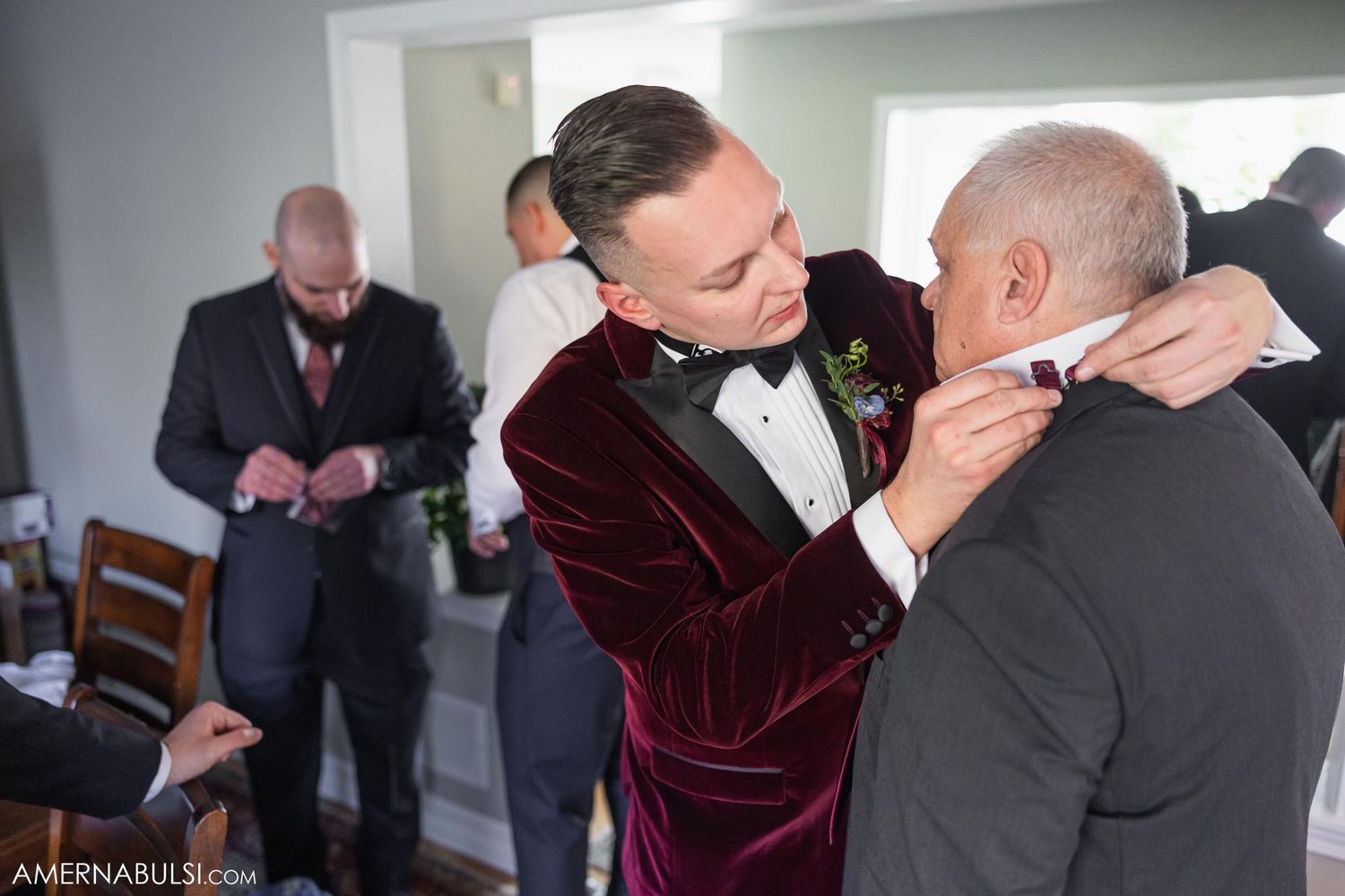 Wedding preparation scene showing groom in burgundy velvet tuxedo helping with a bowtie.