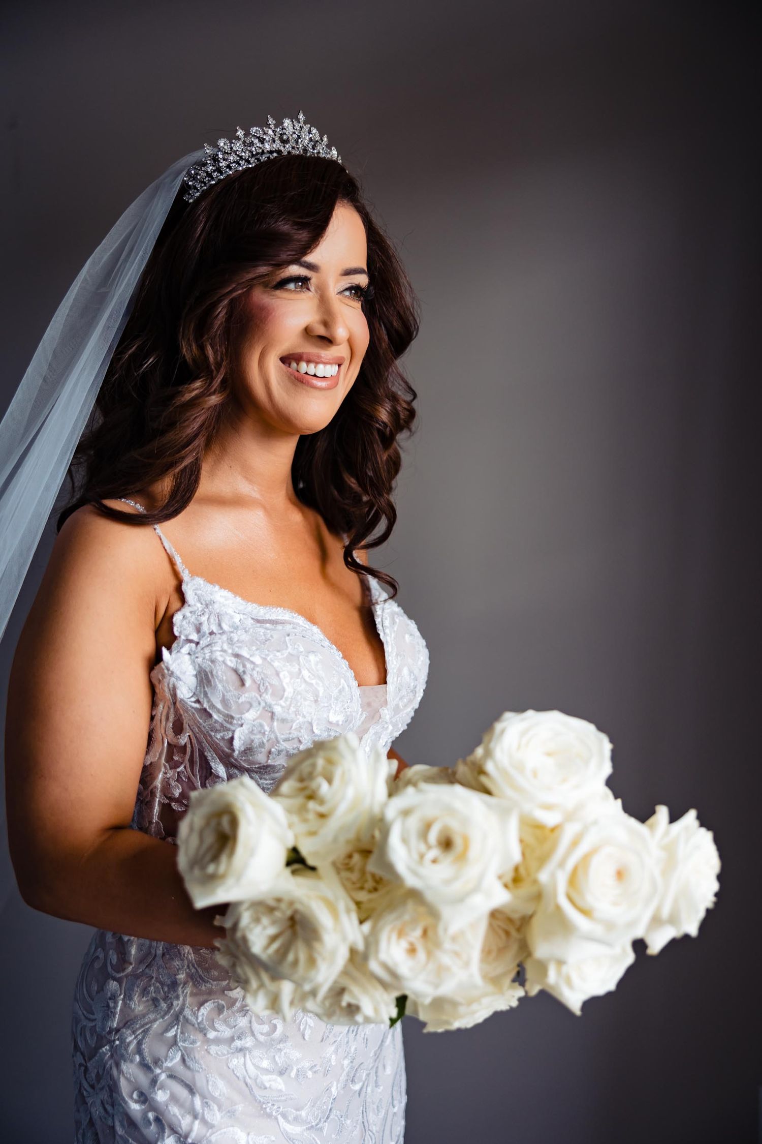 Bride in white lace gown holds cream roses while wearing crystal tiara and veil.